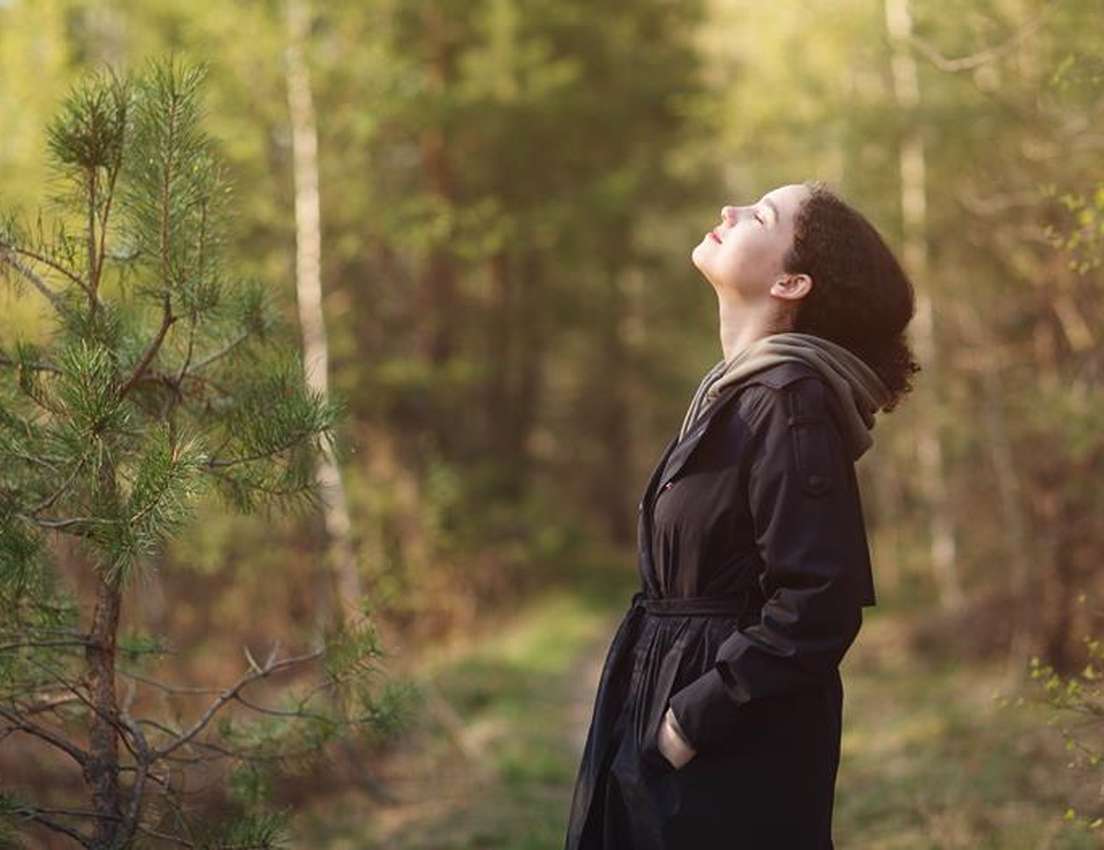 A woman with closed eyes, head tilted up, enjoying the warm light and fresh air in a forest.