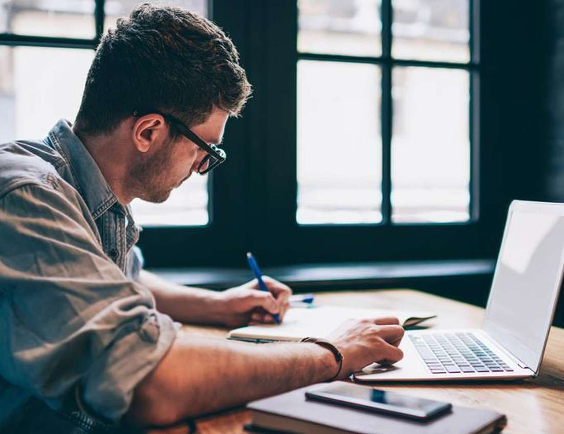Man in glasses and a denim shirt writing in a notebook and typing on a laptop at a wooden desk, with a large window behind hi