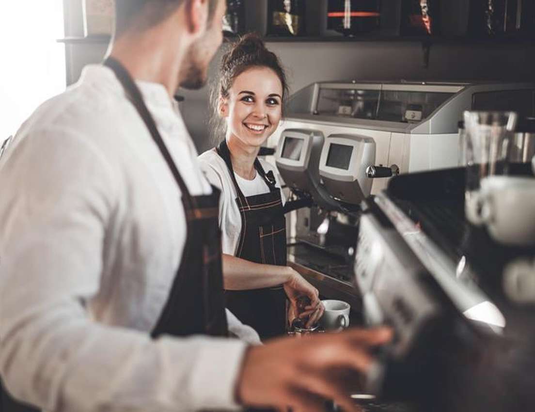 A female barista smiles at a male colleague while making coffee at an espresso machine in a cafe.