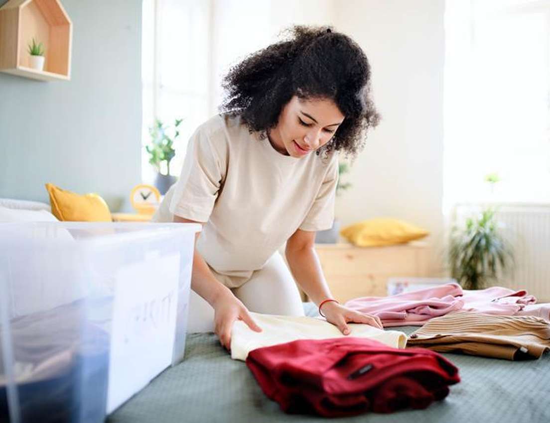 A young woman with curly hair neatly folds a pile of colorful clothes on a bed, placing them next to a clear storage box.