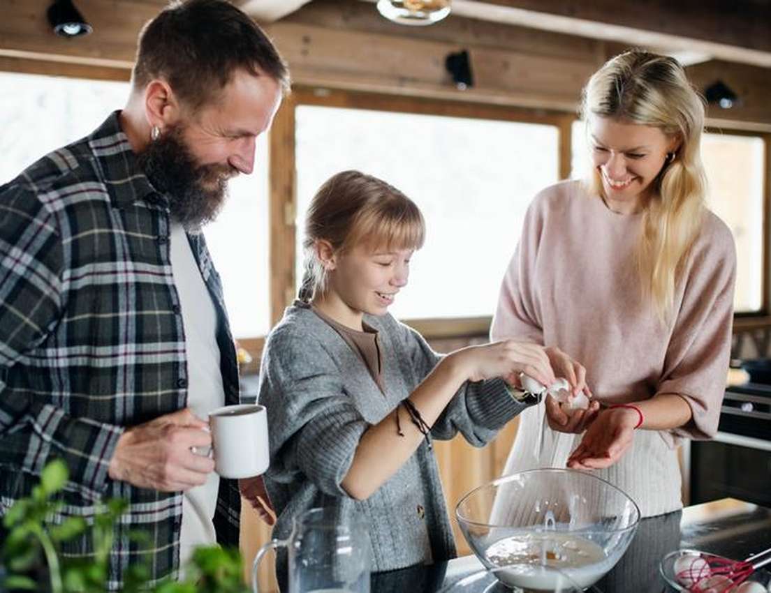 Smiling family cooking in a kitchen. A daughter cracks an egg into a bowl while her parents watch happily.