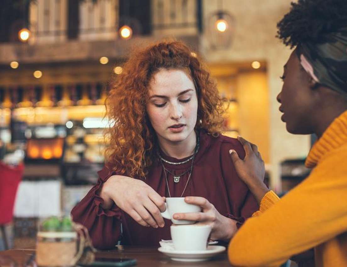 Two women in a cafe. One with red hair holds coffee, looking downcast; the other, in a yellow sweater, comforts her with a ha