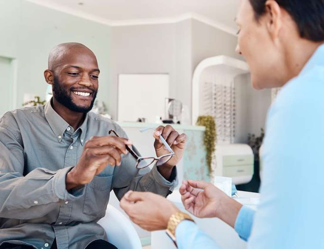 Smiling Black man holding eyeglasses, showing them to an optometrist in an optical store.