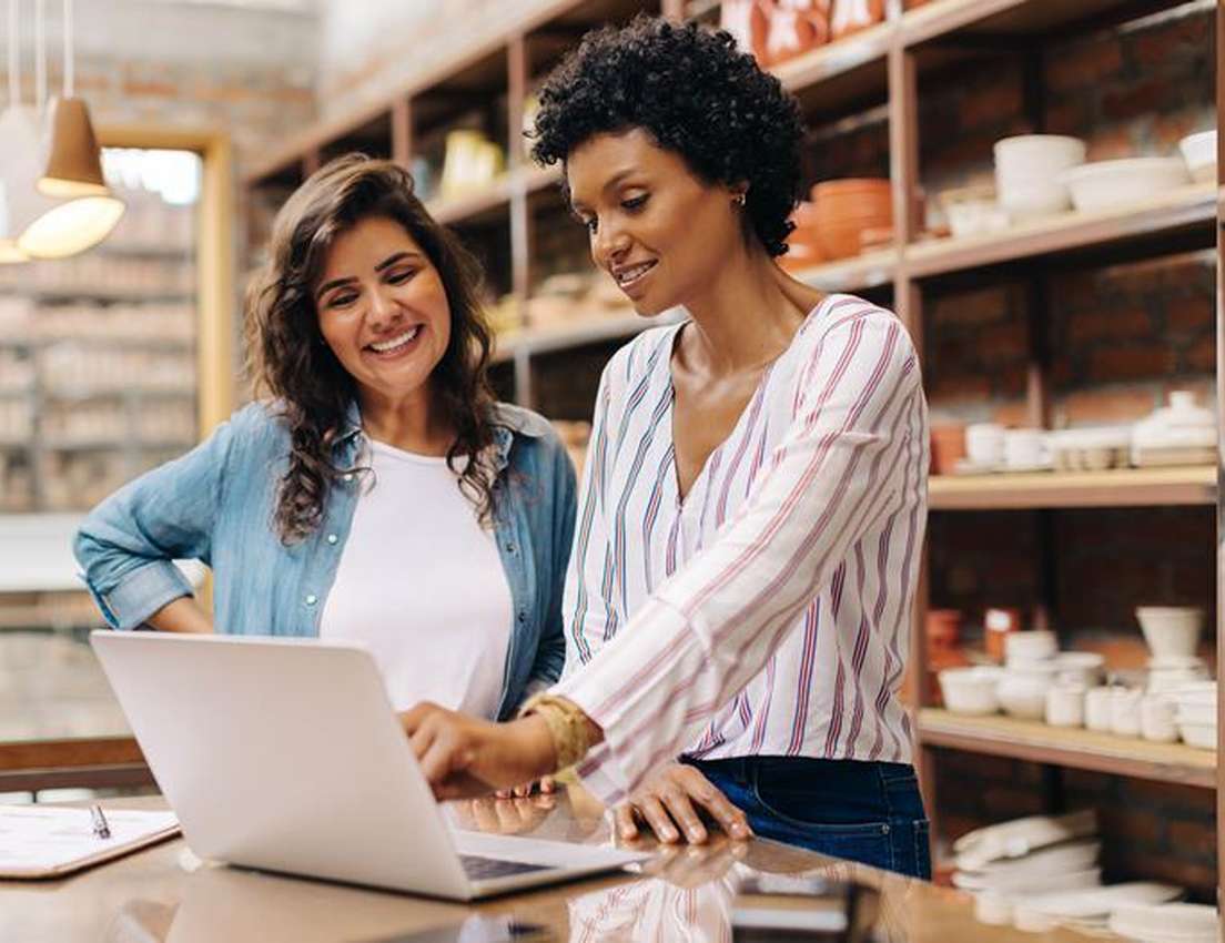 Two women look at a laptop on a counter in a shop filled with pottery shelves; one woman points, the other smiles.