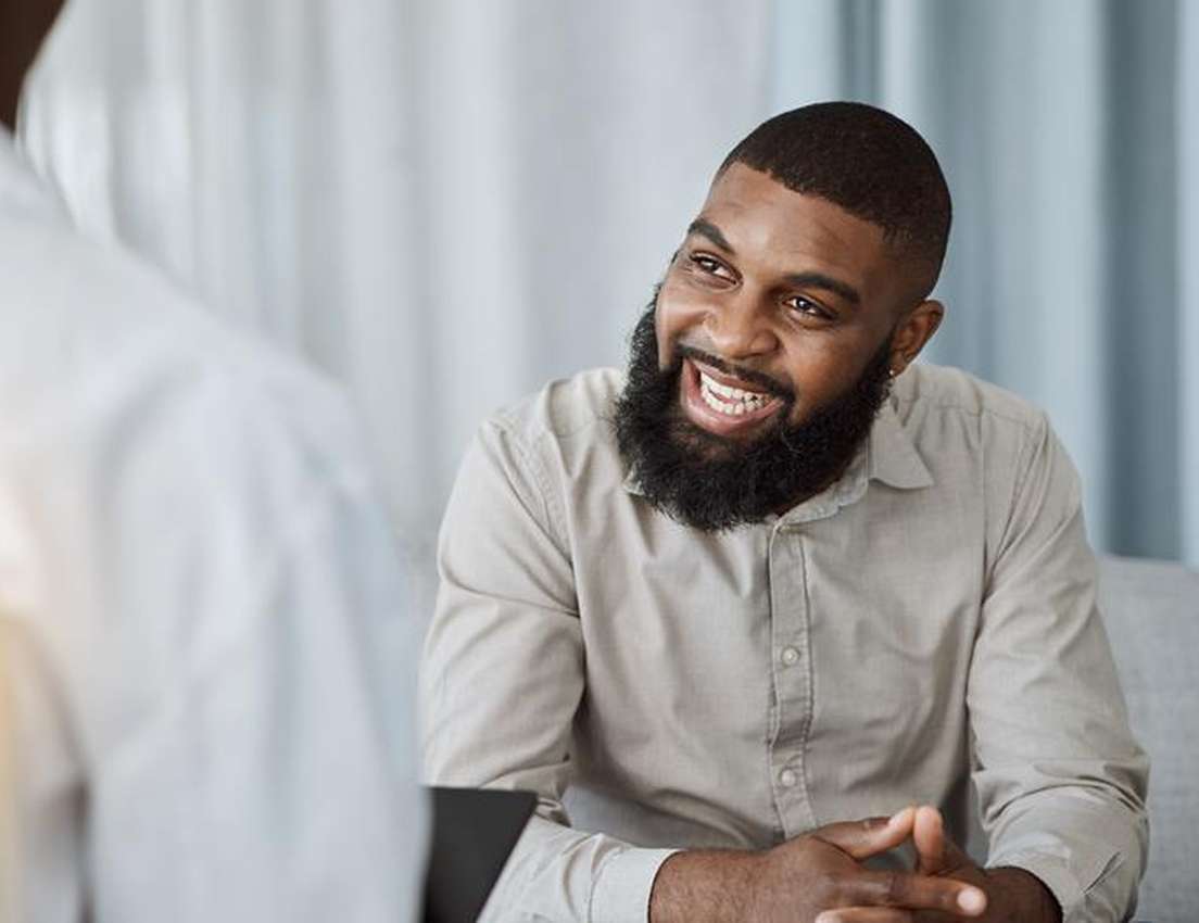 A Black man with a full beard smiles broadly and laughs, looking at a person whose back is to the viewer.