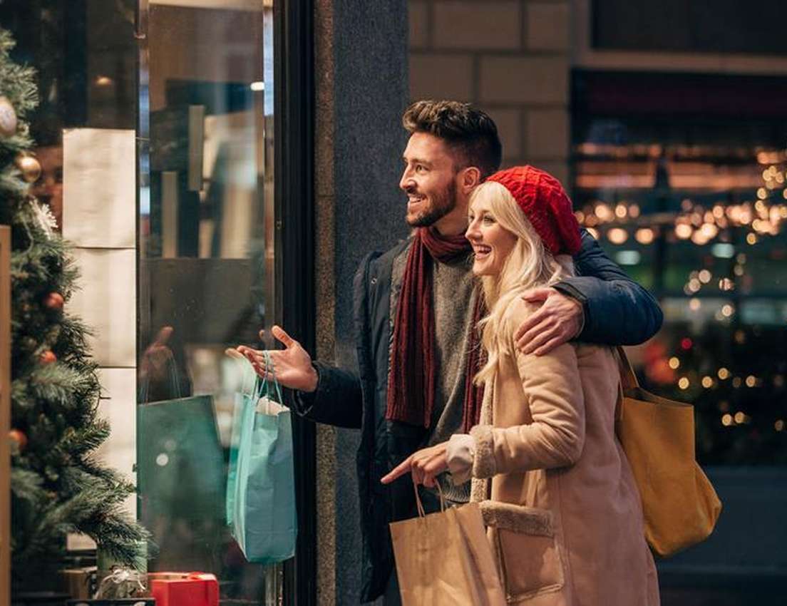 Smiling couple bundled up, window shopping at a festive store with a Christmas tree and blurred lights, holding shopping bags