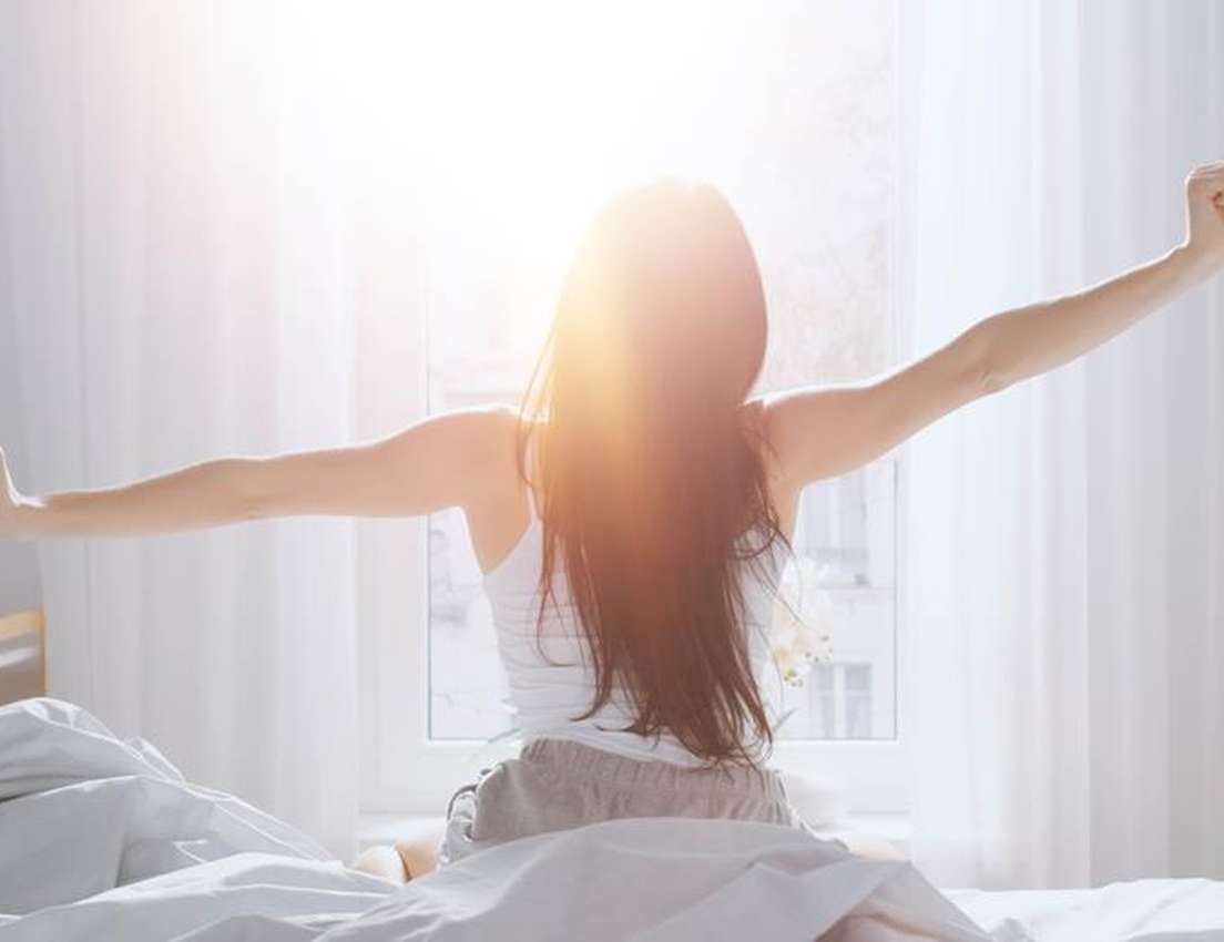 A woman stretches in bed, arms wide, facing a bright, sun-drenched window, her long dark hair illuminated by the morning ligh