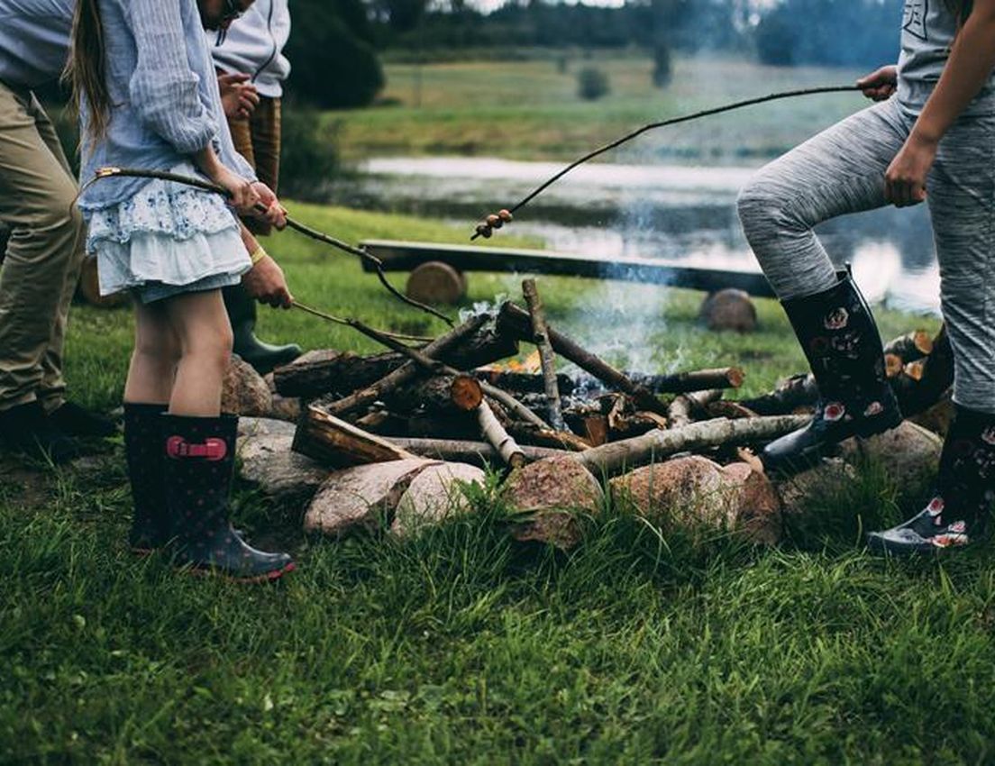 Children roasting marshmallows over a campfire by a lakeside, wearing boots and casual clothes, surrounded by grass and logs.