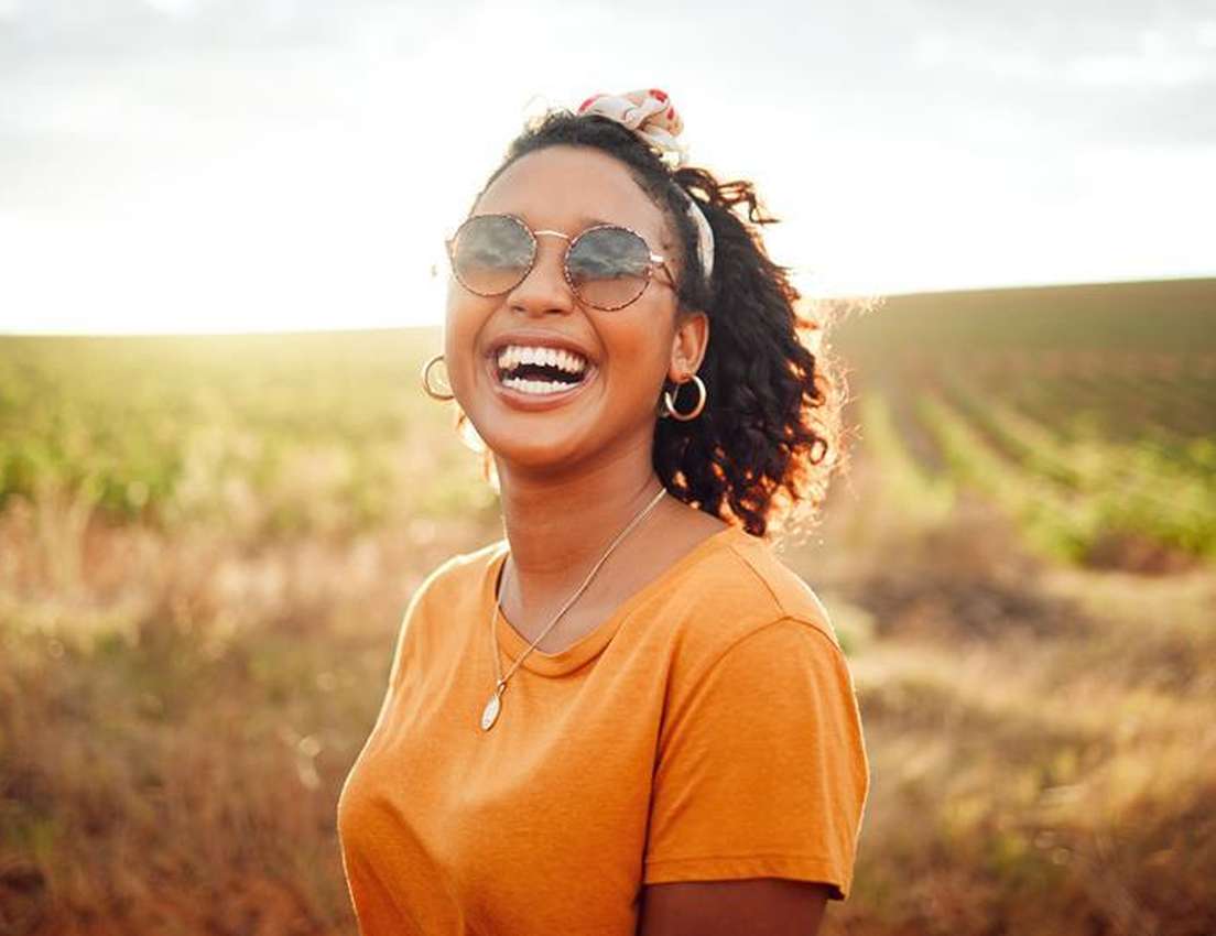 Smiling woman in sunglasses and an orange shirt stands in a sunlit field, with wind-blown hair and leafy plants in the backgr