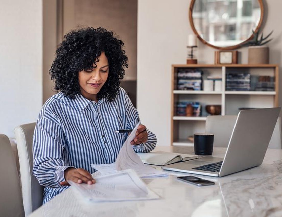 A woman with curly hair reviews documents at a marble table, with a laptop, phone, and mug beside her. A bookshelf is in the