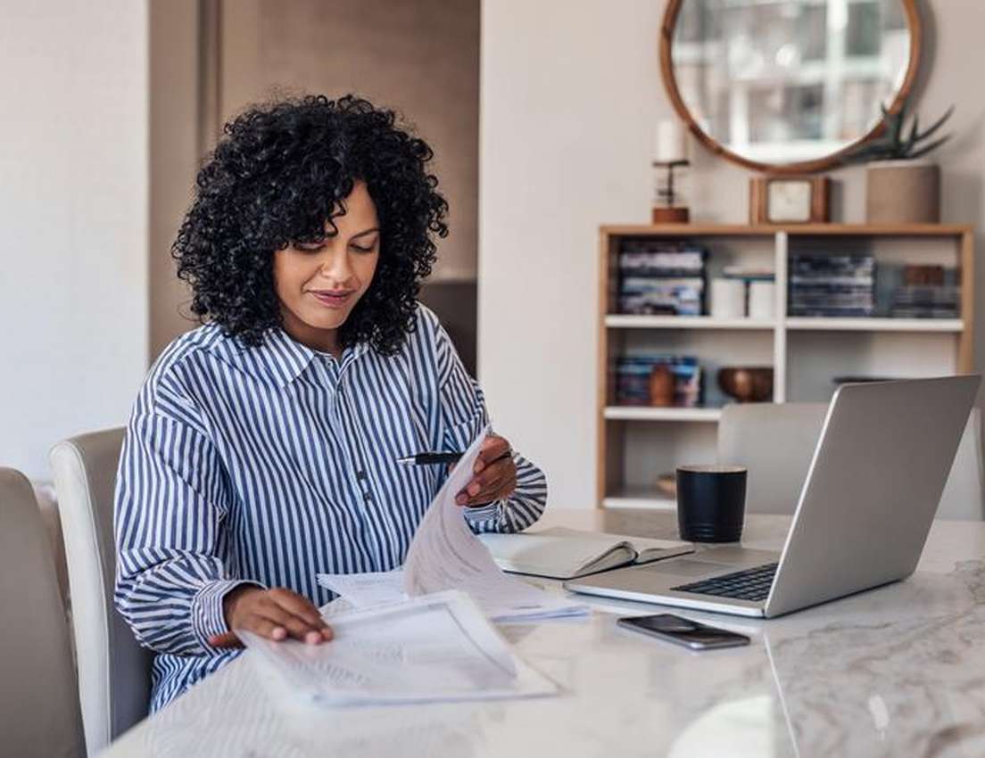 A woman with curly hair reviews documents at a marble table, with a laptop, phone, and mug beside her. A bookshelf is in the 