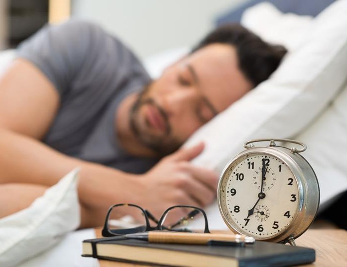 Sleeping man in bed with an alarm clock, glasses, and notebook on the nightstand nearby.