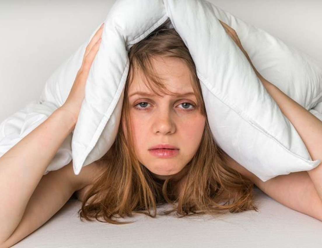 Tired woman in bed covers ears with a pillow, looking exhausted and bothered.