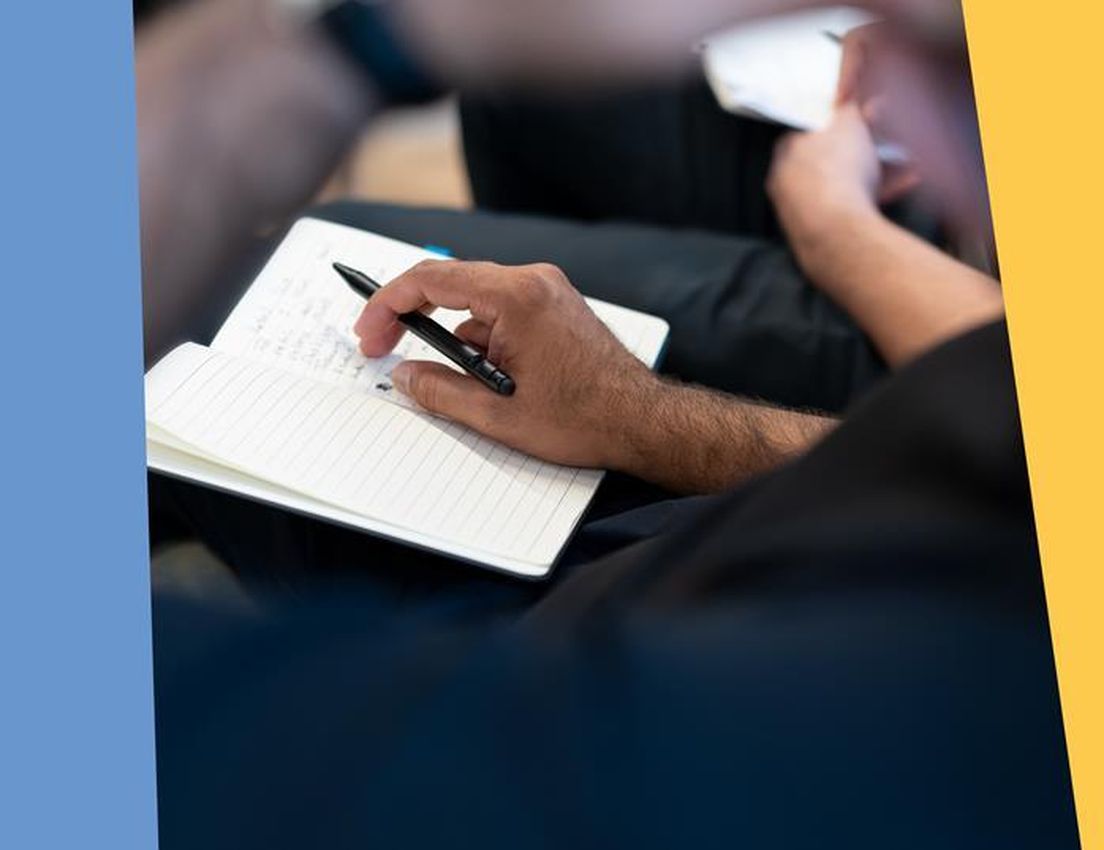 Close-up of a person's hand holding a black pen and writing notes in a lined notebook, with a blue and yellow background.
