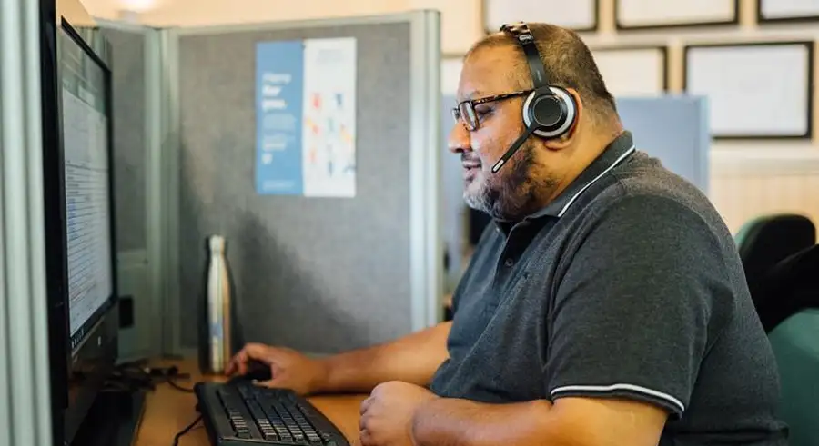 A man with a beard and glasses wears a headset, looking at a computer monitor and using a mouse at a desk in an office cubicl