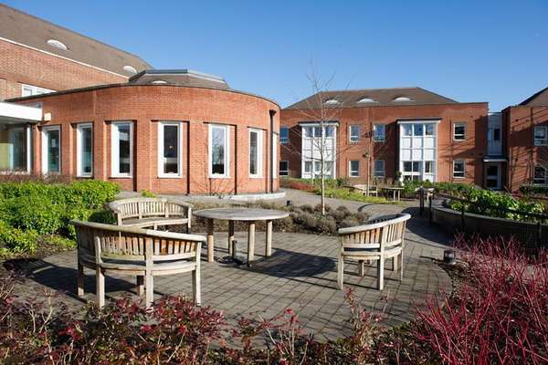 Sunny, paved courtyard with curved wooden benches and a table. Surrounded by green shrubs and red-stemmed plants, with modern brick buildings under a clear blue sky.