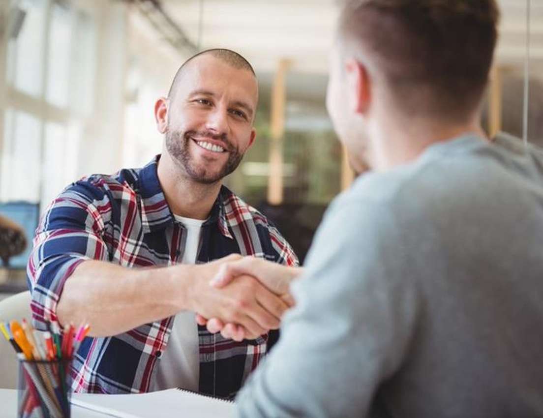 A smiling man in a plaid shirt shakes hands with another man whose back is to the camera, in an office setting.