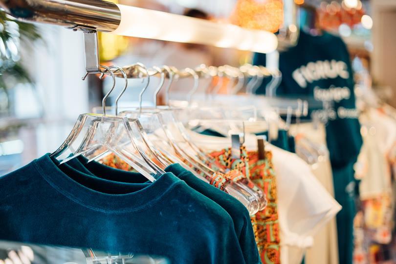 Teal velvet shirts and patterned clothing hang on clear hangers on a metal rack in a brightly lit clothing store.