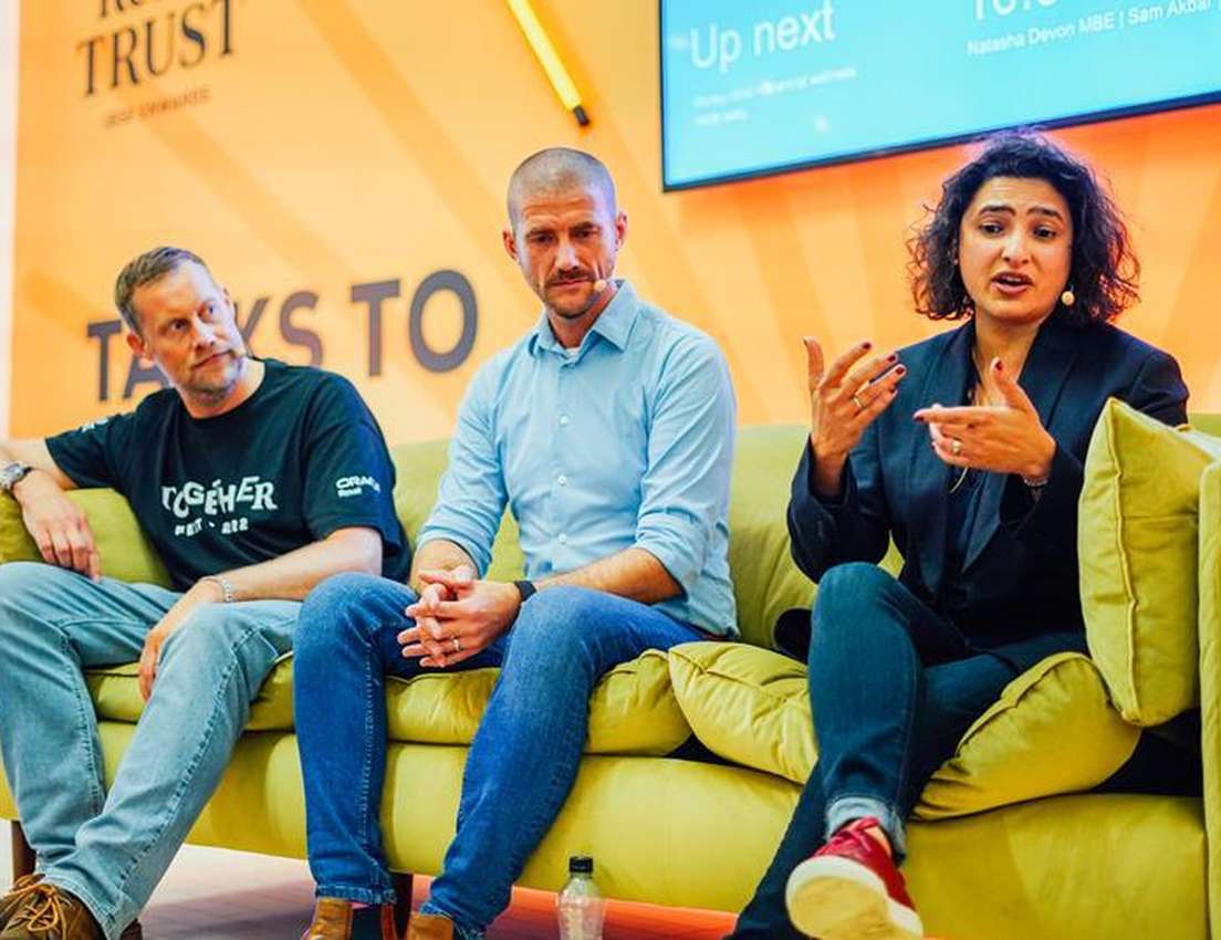 Three people seated on a couch during a panel discussion, with a woman speaking and gesturing, against a vibrant background.