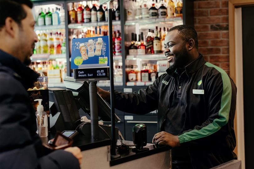Smiling male M&S/BP cashier assists a customer at a checkout counter, showing 'Amount Due £30.00' on screen. Shelves of drinks in background.