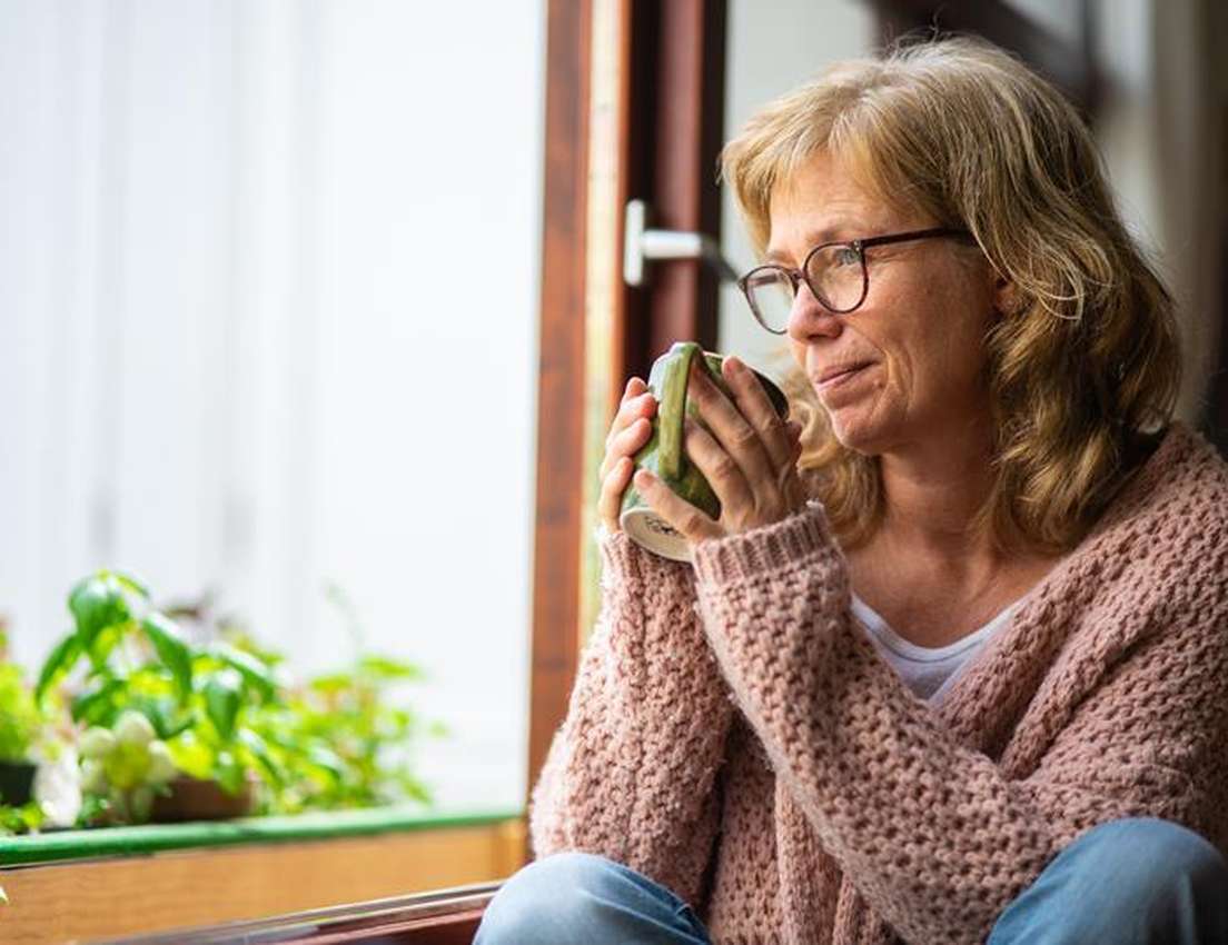 A blonde woman with glasses in a pink cardigan holds a green mug, looking thoughtfully out a window with plants on the sill.