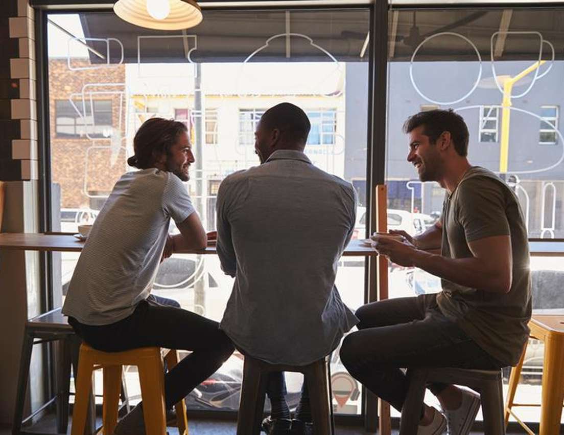 Three friends chat and smile over coffee at a sunlit cafe window.