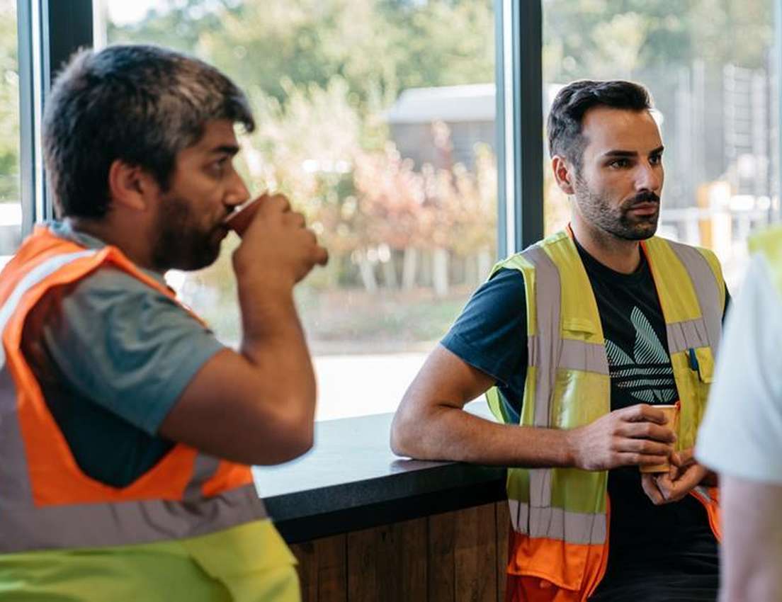 Two men in high-vis vests on a coffee break indoors. One drinks from a cup, the other holds a cup and looks right.