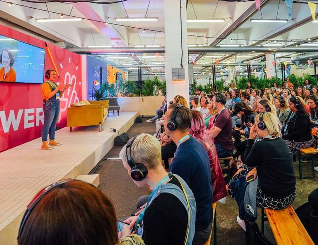 A woman presents on a stage with a screen showing another woman. A large audience wears headphones in a vibrant indoor event 