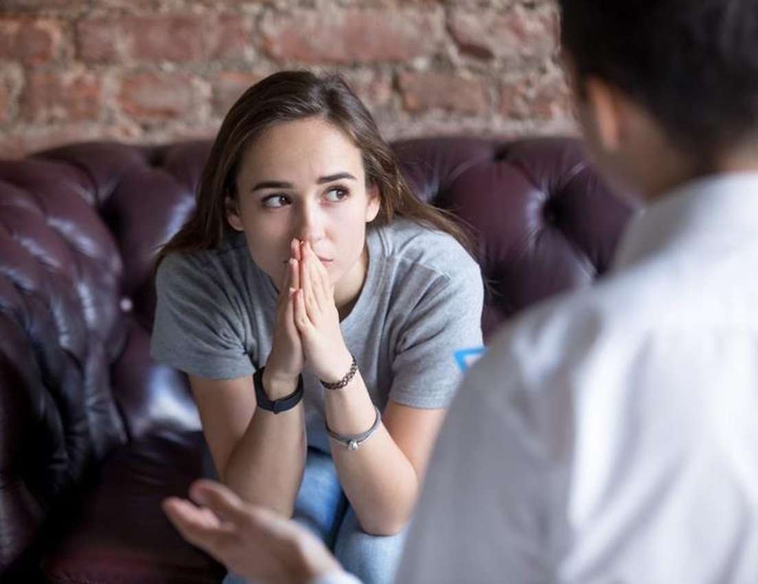 A worried young woman sits on a brown couch, hands clasped, looking away from a person speaking to her.