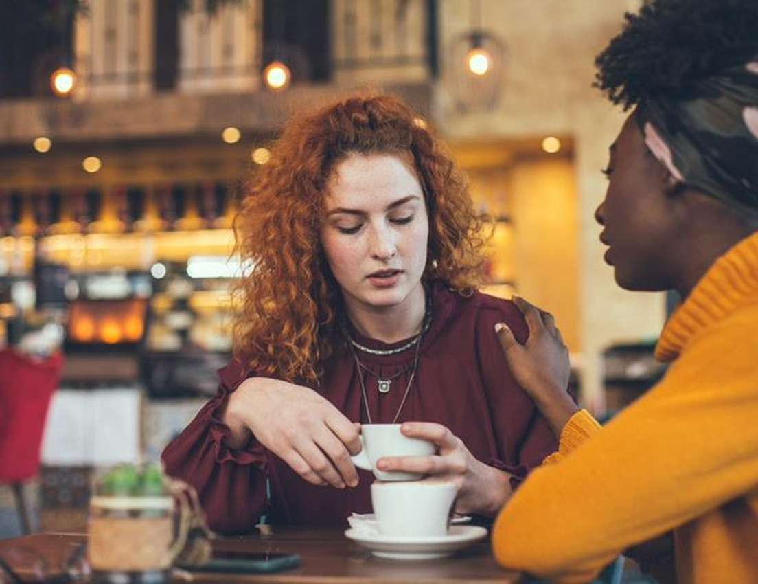 Red-haired woman looks down, holding coffee, as a friend in a yellow sweater comforts her with a hand on her shoulder in a co
