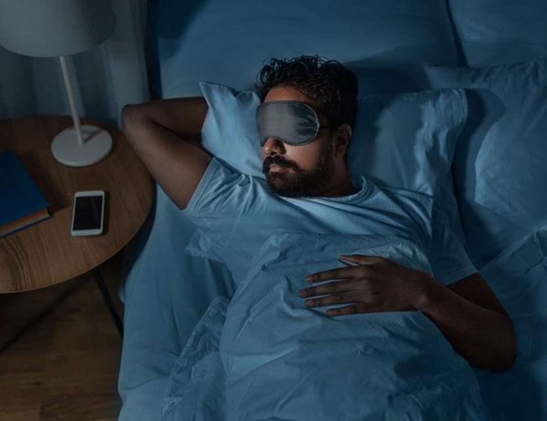 Bearded man in a sleep mask rests in bed at night. Bedside table has a lamp, book, and phone.