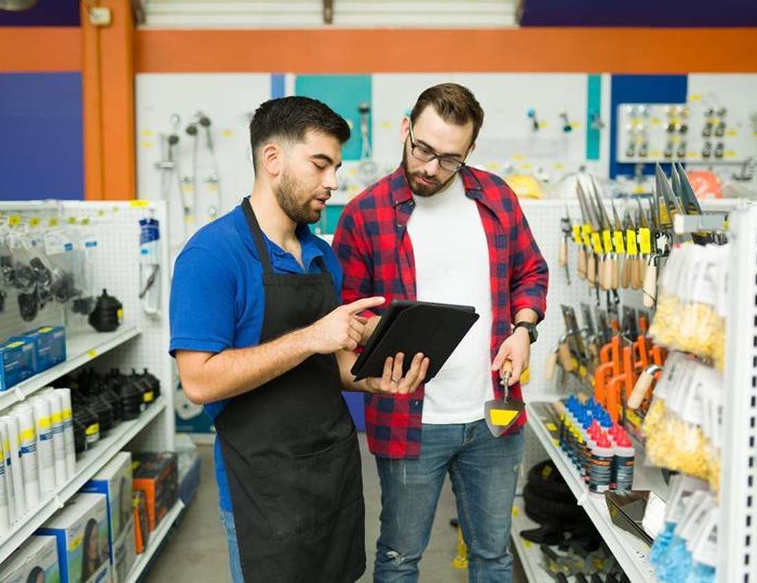 Employee in a blue shirt and apron shows a customer on a tablet in a hardware store filled with tools and supplies.