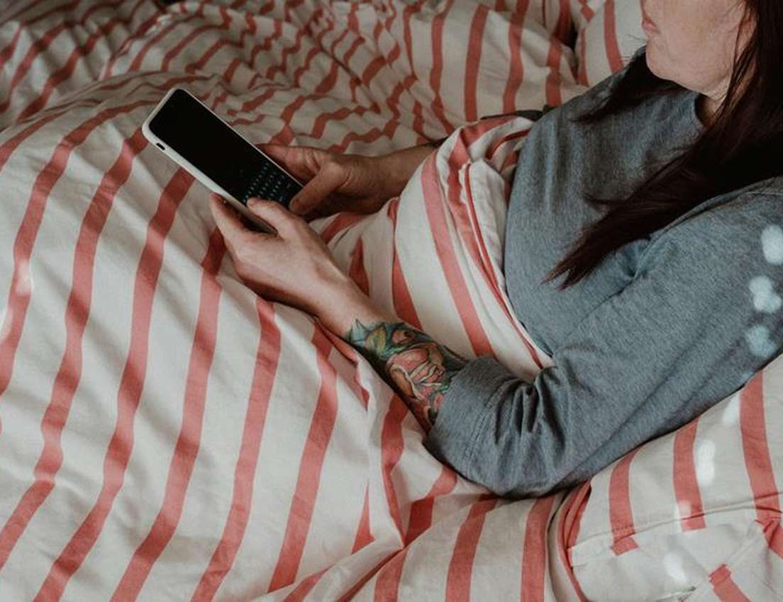 A person lies in bed under a red-striped duvet, holding a white smartphone with a physical keyboard. A tattooed arm is visibl