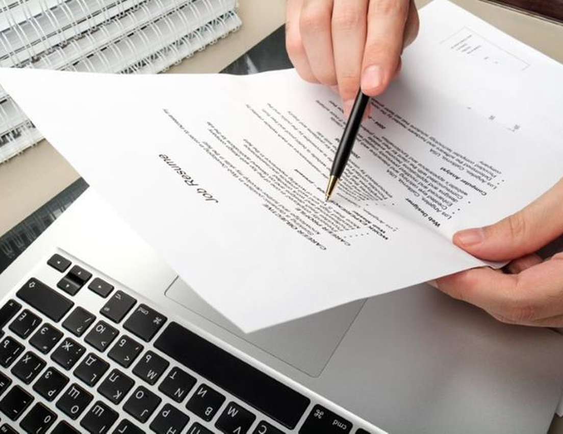 Hands with a pen point at a job resume, placed on a laptop keyboard, with a spiral notebook in the background.