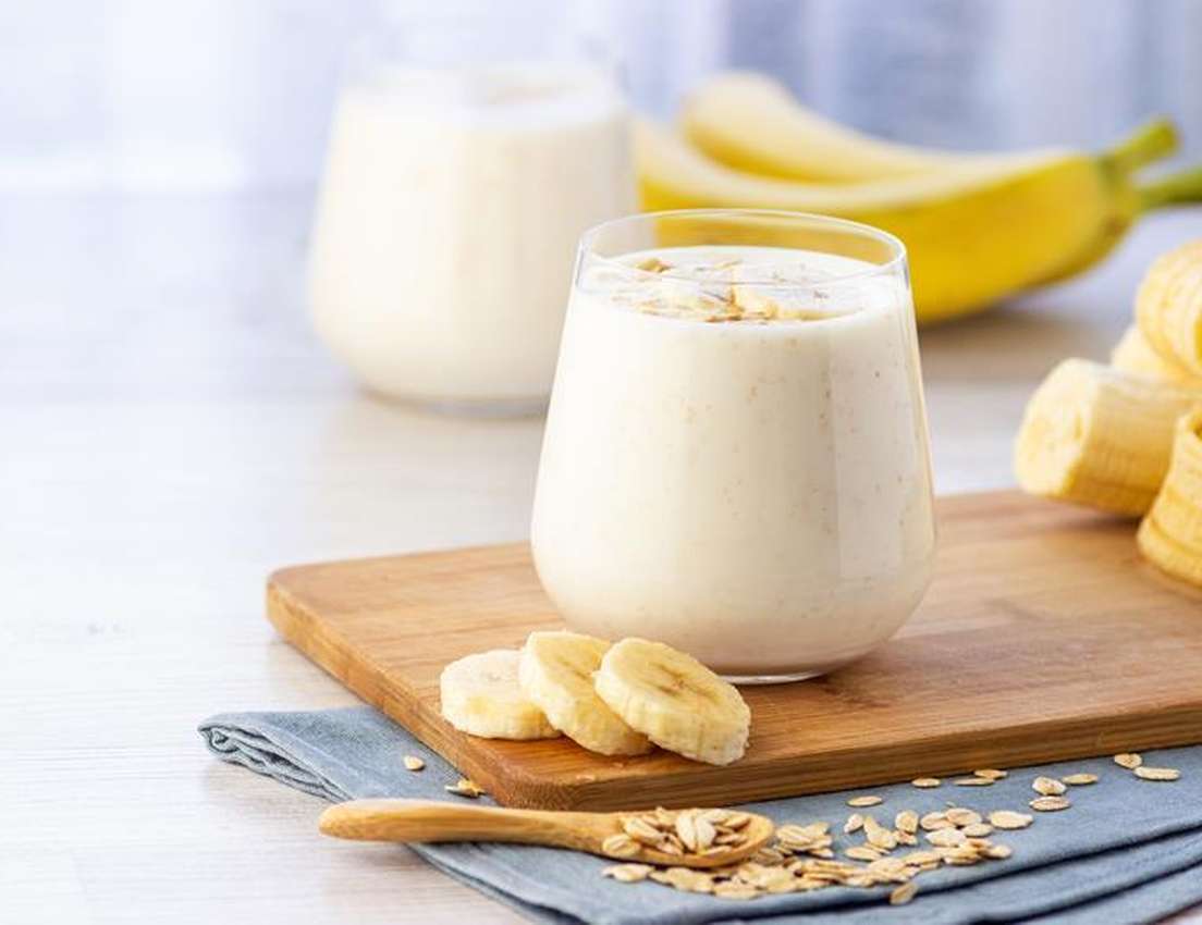 A glass of creamy banana oatmeal smoothie topped with oats, with sliced bananas and a spoon of oats on a wooden board. Banana