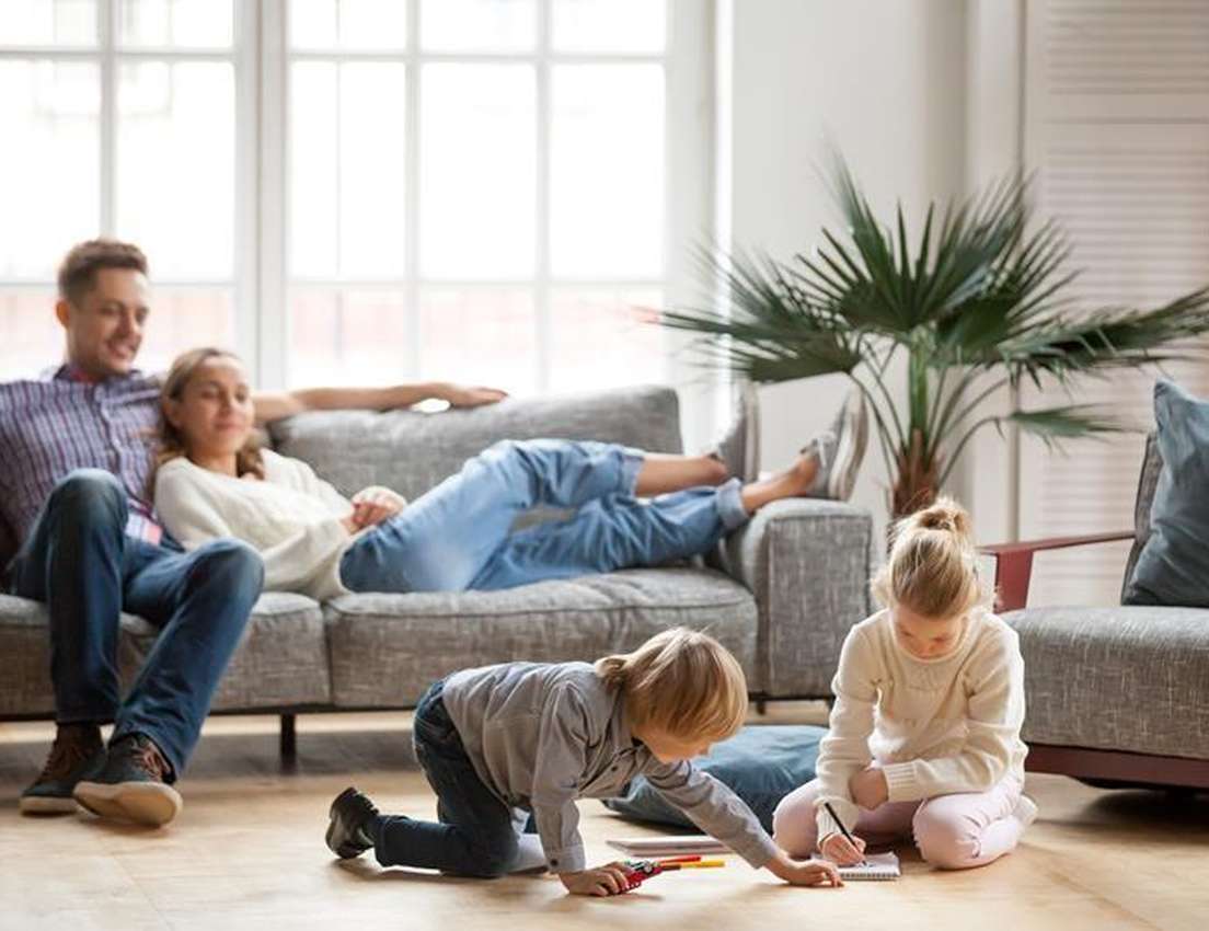 A family in a bright living room: parents relax on the sofa while two young children play and draw on the floor.
