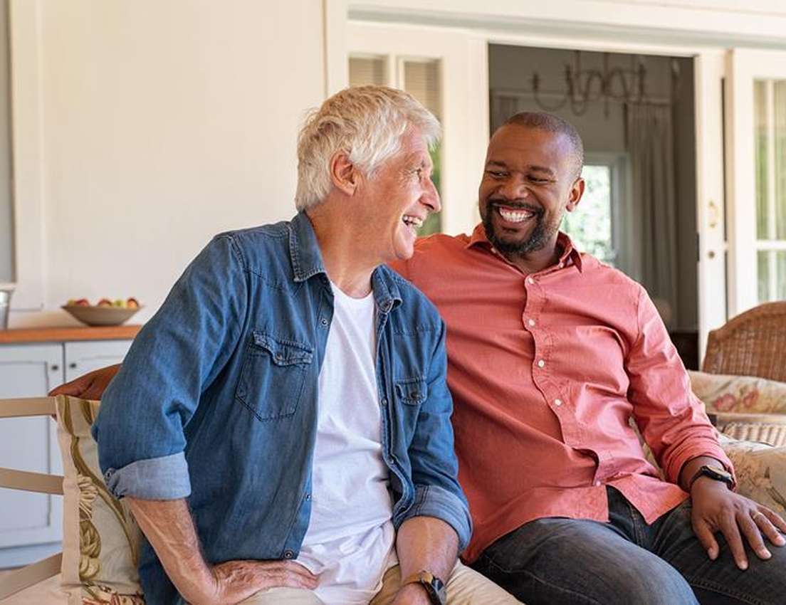 Two men sitting on a porch bench, talking and smiling at each other, creating a warm and friendly atmosphere.