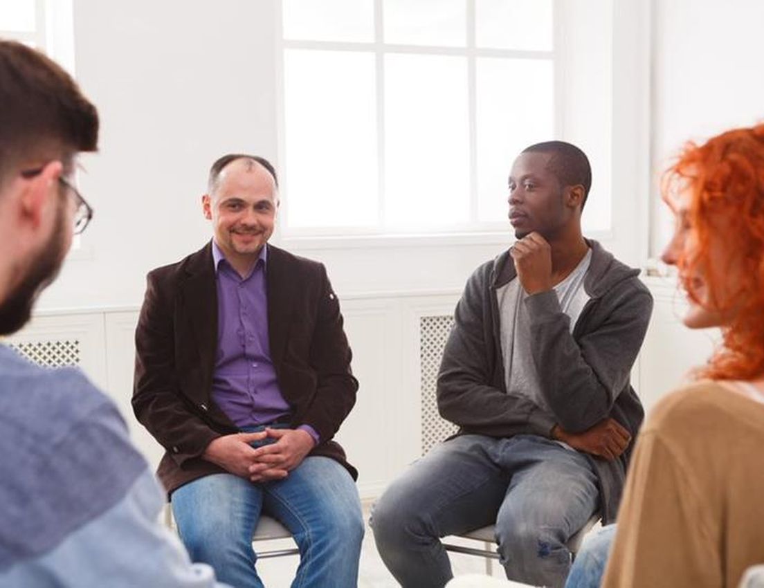 Diverse group of four adults seated in a bright room for a discussion. A man smiles, another listens intently.