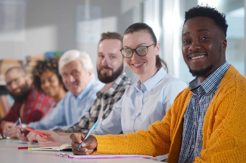 Diverse group of smiling people, including adults and seniors, sit at a table writing. A Black man in a yellow cardigan looks at the camera.