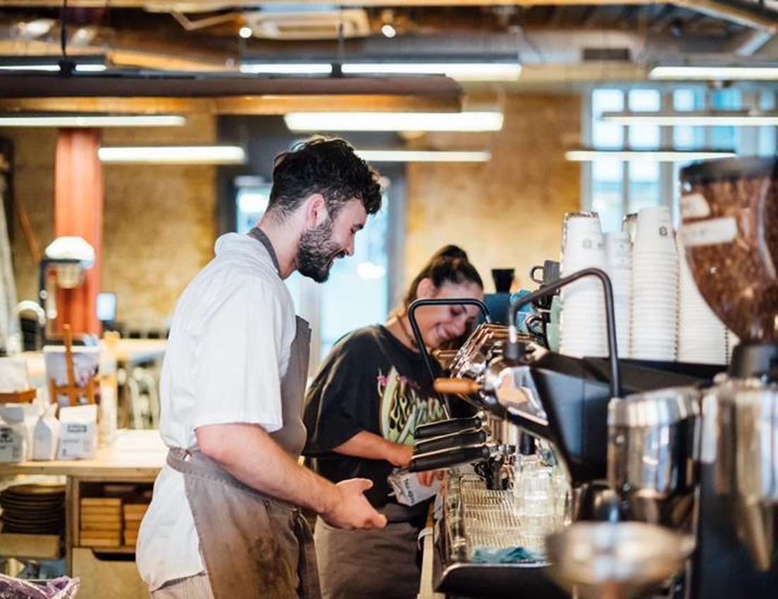 Two smiling baristas, a man and a woman, work together at a coffee shop counter with machines, paper cups, and coffee beans.