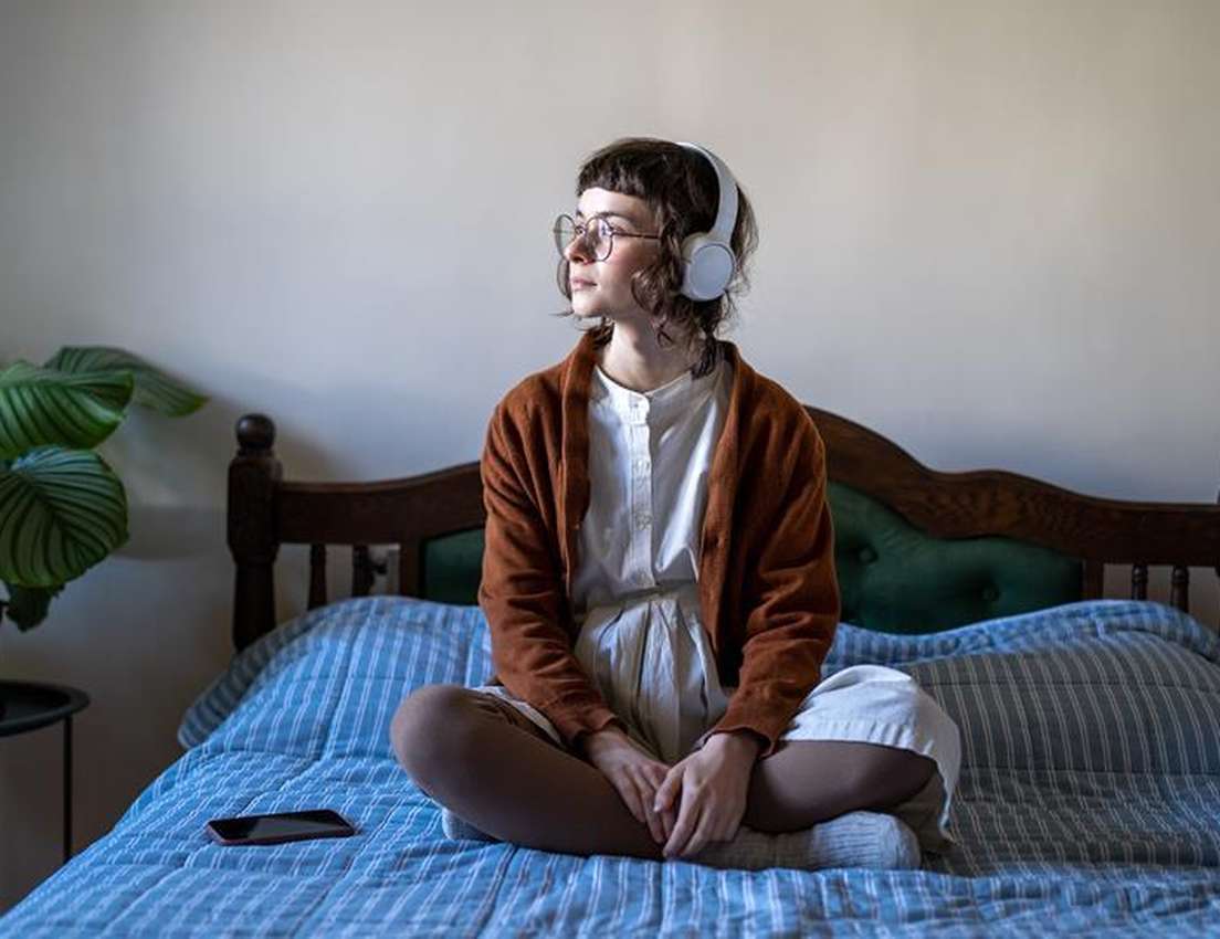 Person sits cross-legged on a bed, wearing headphones and glasses, looking thoughtful. A smartphone and potted plant are near