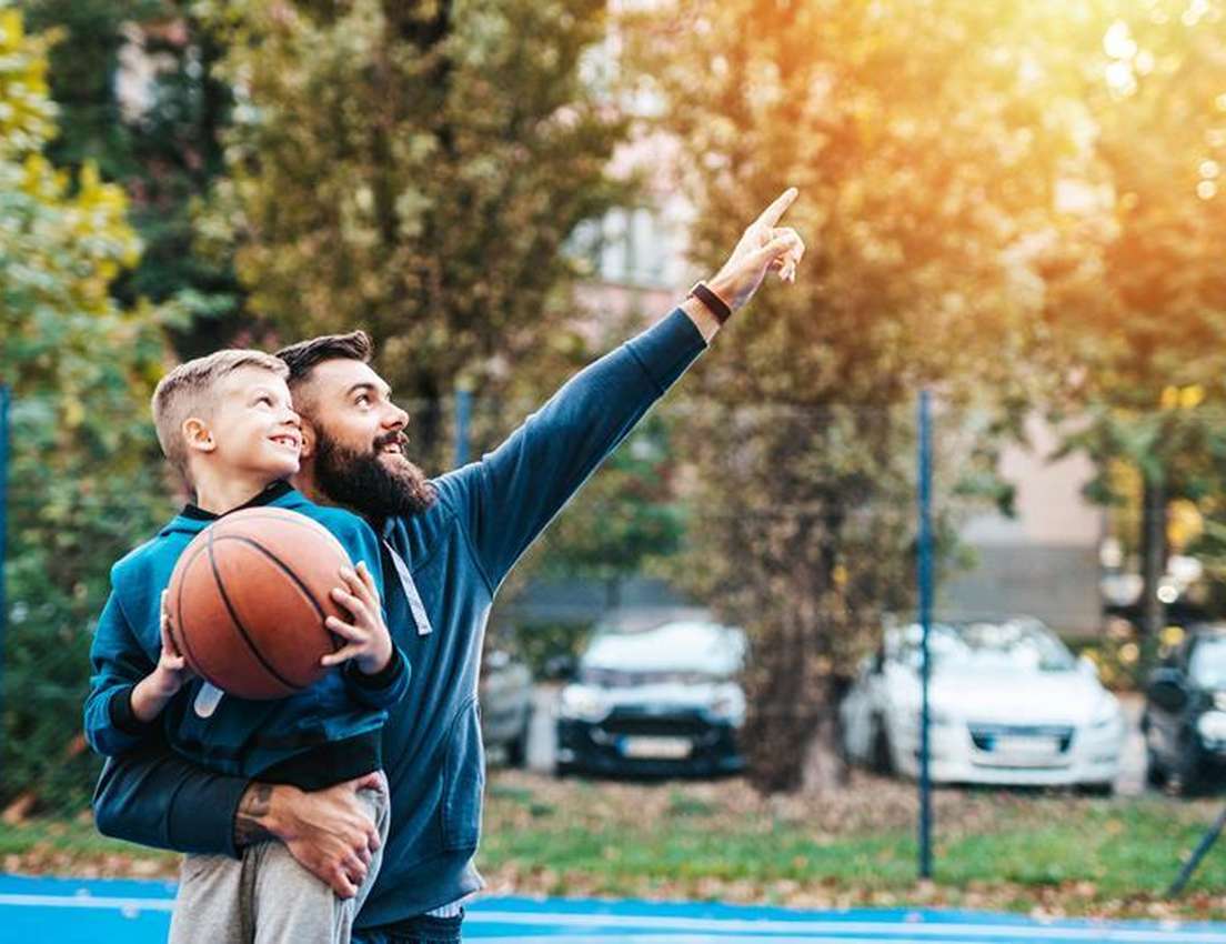 Smiling father and son on a basketball court. The dad holds the boy, who has a basketball. Both look up as the dad points int