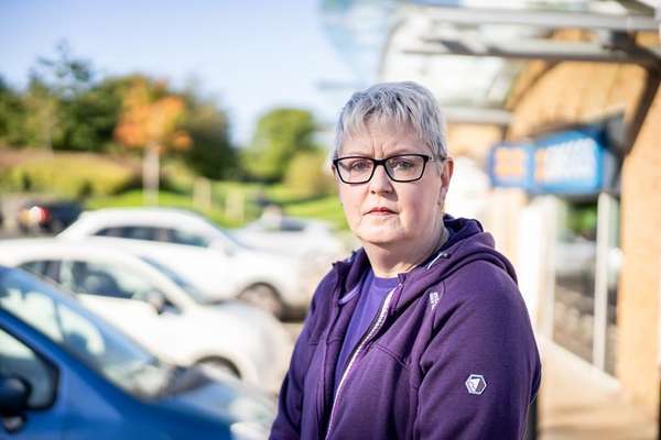 A serious-looking woman with short grey hair and glasses in a purple hoodie stands in a sunny parking lot with cars behind her.