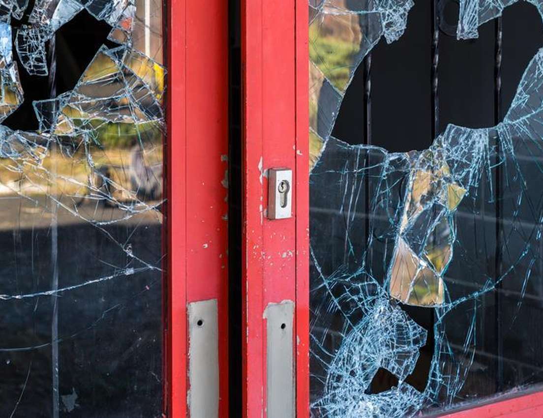 Close-up of a red-framed glass door with shattered glass, showing cracks and missing pieces around a visible keyhole.
