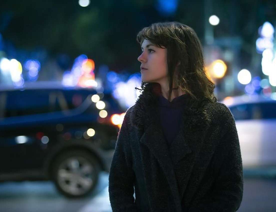 Woman in a dark coat stands on a city street at night, surrounded by blurred car lights and colorful bokeh in the background.