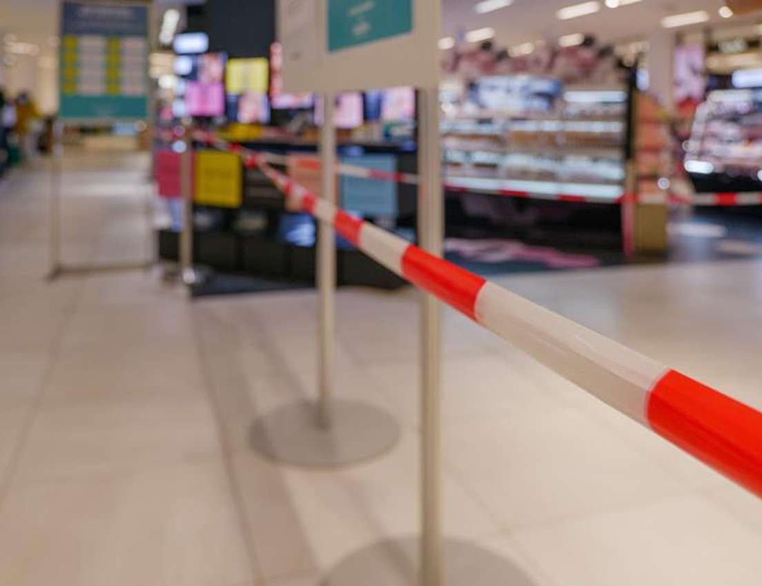 A red and white striped barrier tape blocks off an area in a brightly lit store, with blurred display shelves and people in t