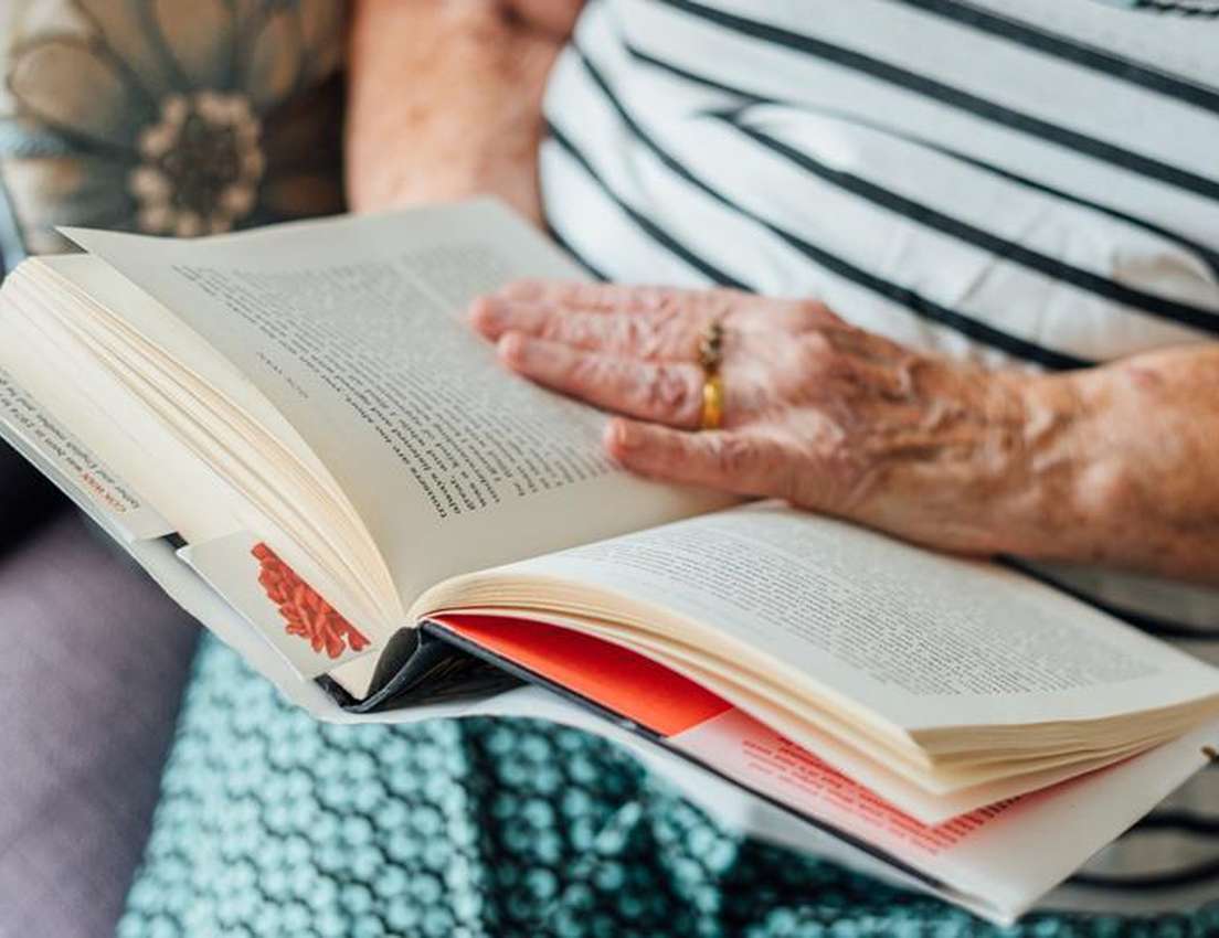 An elderly person wearing striped clothing reads a book, with one hand resting on the pages, seated on a patterned cushion.