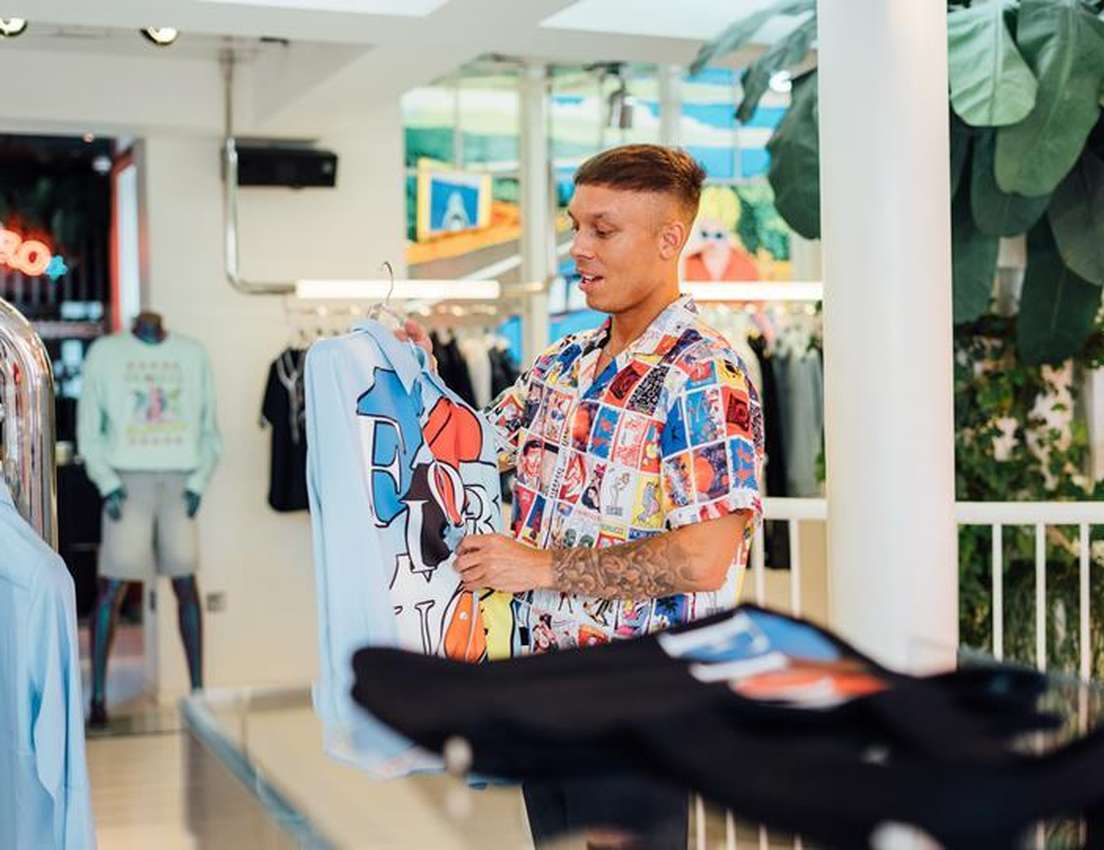 A man in a comic-strip shirt smiles while holding a light blue graphic t-shirt in a stylish boutique.