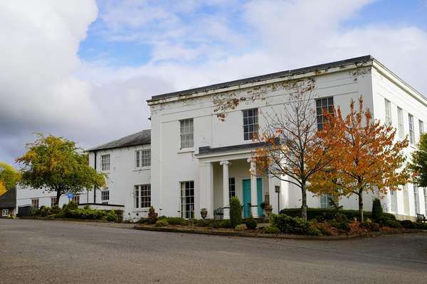 White classical building with turquoise doors and peeling paint, surrounded by autumn trees under a cloudy sky.