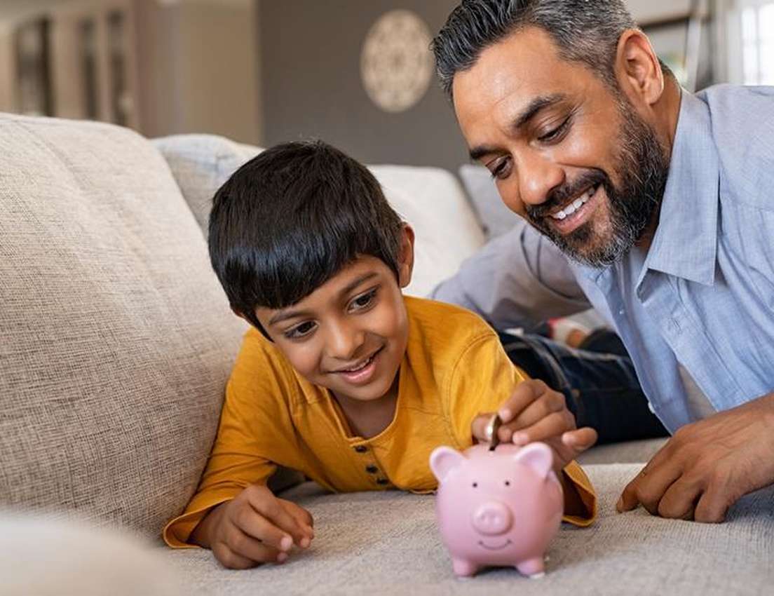 A smiling father watches his young son put a coin into a pink piggy bank on a couch.